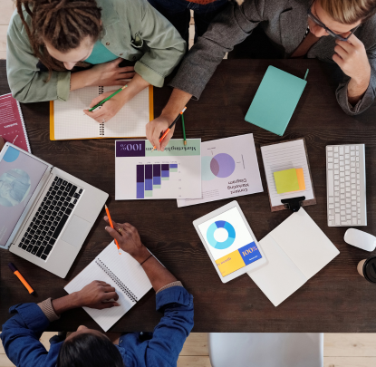 Tavolo di  lavoro visto dall'alto con tre persone, fogli con grafici e pc