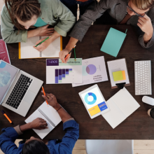 Tavolo di  lavoro visto dall'alto con tre persone, fogli con grafici e pc